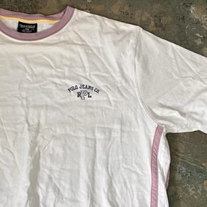 May include: A white short-sleeve t-shirt with a mauve collar and sleeve trim. The chest features the embroidered text Polo Jeans Co. and the letters RPL. A mauve vertical stripe runs down the side of the shirt, and a black label is at the neck.