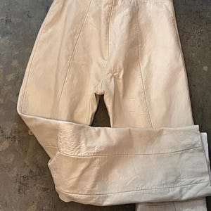 May include: Off-white leather trousers featuring a high-waisted design with vertical seam detailing running down the front of each leg. The pants have a smooth texture, a side zipper closure, and a straight-leg cut with a horizontal seam above the hem.