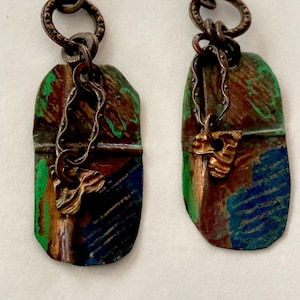 May include: A pair of handmade earrings with a rustic, artistic design. Each earring features a rectangular, textured pendant with green, blue, and brown paint. A small, gold-colored charm hangs from a chain attached to the pendant.