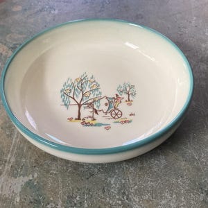 May include: A round, off-white ceramic bowl with a teal rim and a decorative scene. The scene depicts a horse-drawn carriage, trees, and a well. The bowl has a slightly raised edge and is suitable for serving food.