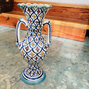 May include: A white ceramic vase with blue, green, yellow, and red geometric patterns. The vase has two handles and a scalloped rim.