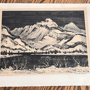 May include: A black and white print on paper depicting a mountain range with snow-covered peaks, a dark lake, and a foreground of bare trees and shrubs. The artwork features a signature in the bottom right corner and is set on a light border.