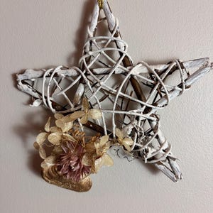 Puede incluir: Decoración en forma de estrella, hecha de ramas trenzadas pintadas de blanco, con una cinta dorada para colgar. Adornada con flores secas y un elemento decorativo dorado, creando una estética rústica y festiva. La estrella mide unos 25 cm de diámetro.