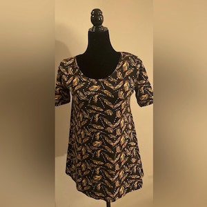 May include: Black short-sleeved top with a scoop neckline. The fabric is patterned with colourful feathers in yellow, blue, and orange. The feathers are arranged in a repeating pattern across the material.