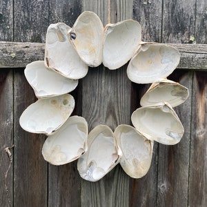 Peut inclure: Une couronne faite de coquilles de palourdes blanches, disposées en cercle et accrochée à une clôture en bois.