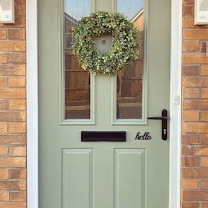 May include: A light green front door with a black door handle and a black letterbox. A green wreath hangs on the door. The word "hello" is written in black lettering on the door.