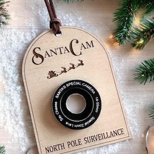 May include: A wooden Christmas bauble shaped like a camera lens with the text "SantaCam" and "North Pole Surveillance" printed on it. The lens has the text "Santa's Special Camera" and "Special Zoom Lens 14-55mm" printed around it.