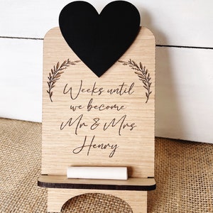 May include: Wooden countdown sign with a black heart chalkboard and a piece of chalk. The sign says "Weeks until we become Mr & Mrs Henry".