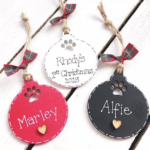 May include: Three personalised Christmas baubles with a paw print cutout. The baubles are red, white and black with white lettering. The text on the baubles reads "Marley", "Rhody's 1st Christmas 2018", and "Alfie".
