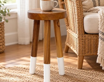 May include: A small, round wooden side table with three legs, each featuring a white-dipped base. A light-colored ceramic mug sits on the flat, circular top surface of the table, which displays a natural wood grain finish throughout.