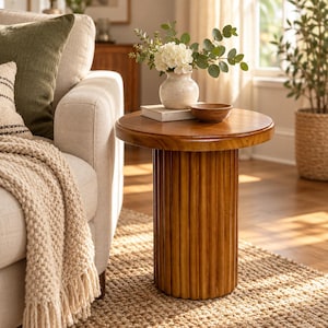May include: A round wooden side table with a fluted, cylindrical base. The table features a smooth, polished top surface. On the table sits a small, speckled off-white ceramic vase containing white flowers and green eucalyptus leaves.