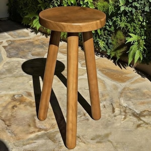 May include: A wooden stool with a round seat and three angled legs. The stool is made of light brown wood and is set against a backdrop of greenery and stone. The stool's design is simple and functional.