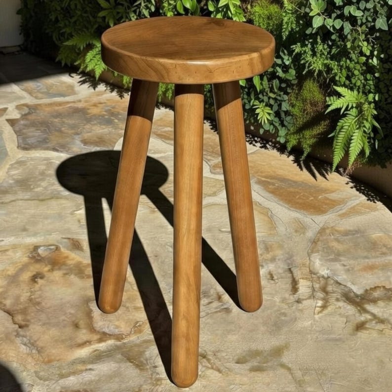May include: A wooden stool with a round seat and three angled legs. The stool is made of light brown wood and is set against a backdrop of greenery and stone. The stool's design is simple and functional.