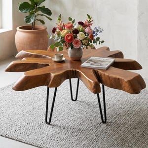 May include: A coffee table featuring a natural wood slab top with an irregular, organic edge and three black metal hairpin legs. A ceramic vase with a colorful floral arrangement, a small cup, and a book titled Slow Living sit on top.