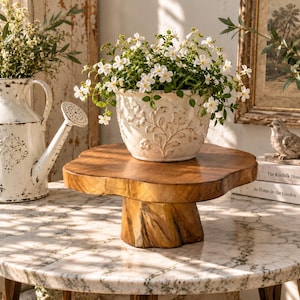 May include: A round wooden pedestal stand with a natural edge and a thick central base, supporting a white ceramic planter featuring an embossed floral pattern and filled with small white flowers, positioned on a white marble surface.