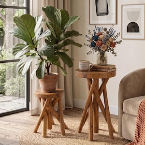May include: Two round wooden side tables with crossed leg bases. The shorter table holds a large green fiddle leaf fig plant in a terracotta pot. The taller table holds a ceramic mug with the text Morning Brew and a bouquet of dried flowers.