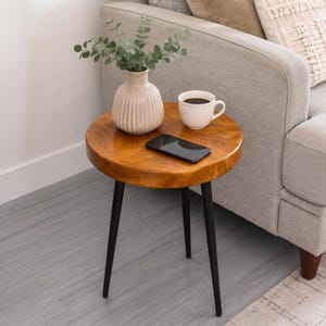 May include: A round wooden side table with three slender black metal legs. On the tabletop sits a ribbed cream-colored ceramic vase containing green eucalyptus stems, a white ribbed ceramic mug filled with black coffee, and a black smartphone.