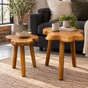 May include: Two natural wood side tables with organic, irregular-shaped tops and three sturdy cylindrical legs. The smaller table holds a shallow ceramic bowl, while the larger table supports a ceramic mug and a small potted plant.
