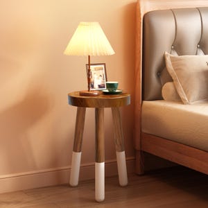 May include: A small, round wooden side table with three legs, each tipped with white paint. A lamp with a pleated shade and a framed photo sit on top, along with a small green teacup and saucer. The table is next to a bed.
