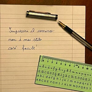 Puede incluir: Una pluma estilográfica y un reposaplumas sobre papel rayado con escritura cursiva italiana. Una regla verde con ejemplos del alfabeto cursivo está en la esquina inferior derecha. El texto en el papel dice "Imparare il corsivo non è mai stato così facile!"