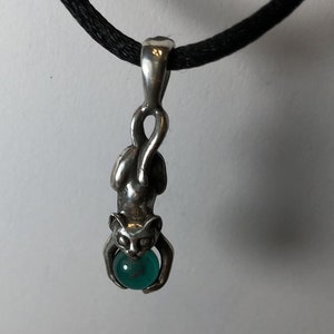 Puede incluir: Un collar con un colgante de plata en un cordón negro. El colgante presenta una figura de gato de plata que sostiene una piedra preciosa de color turquesa.