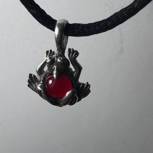 Puede incluir: Un colgante de rana de plata con una piedra roja en forma de corazón en su vientre, colgando de un cordón negro.