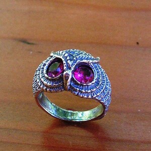 Peut inclure: Bague argentée en forme de hibou, ornée de deux pierres magenta rondes en guise d'yeux. La tête du hibou présente des motifs texturés, et l'anneau est lisse. Posée sur une surface en bois.