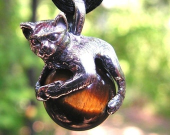 Peut inclure: Un pendentif en argent en forme de chat avec une pierre précieuse en œil de tigre. Le chat tient la pierre précieuse dans ses pattes. Le pendentif est sur un cordon noir.