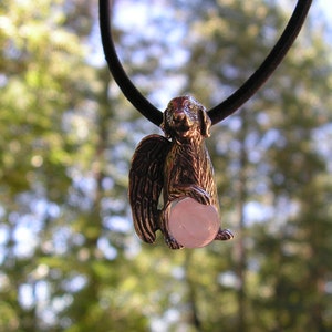 May include: A silver pendant necklace featuring a detailed sculpture of a dog with wings holding a pink gemstone. The dog is facing forward with its head tilted slightly to the side.