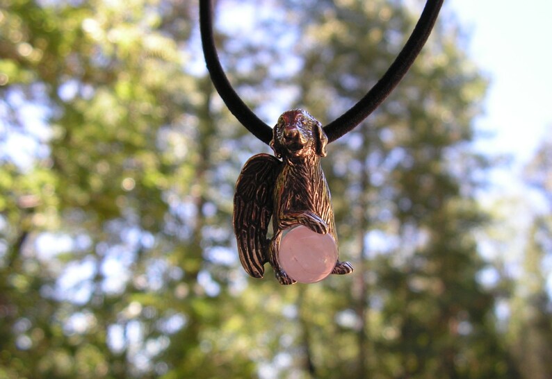 May include: A silver pendant necklace featuring a detailed sculpture of a dog with wings holding a pink gemstone. The dog is facing forward with its head tilted slightly to the side.