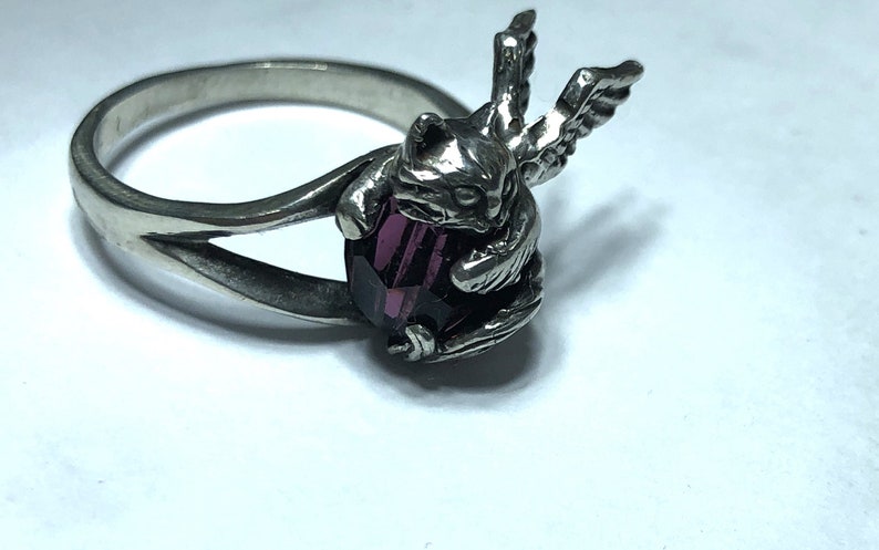 May include: A silver ring with a winged creature holding a purple gemstone. The ring has a split band design.