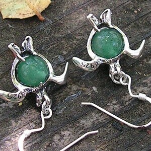 Pu&ograve; includere: Un paio di orecchini d'argento con accenti di pietre preziose verdi. Ogni orecchino presenta un design di tartaruga dettagliato con una pietra verde rotonda al centro. Gli orecchini hanno ganci d'argento.