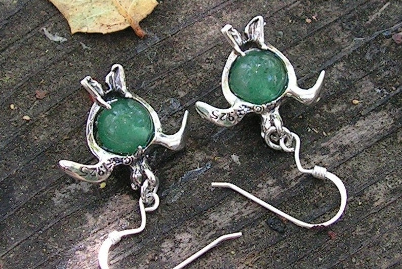Pu&ograve; includere: Un paio di orecchini d'argento con accenti di pietre preziose verdi. Ogni orecchino presenta un design di tartaruga dettagliato con una pietra verde rotonda al centro. Gli orecchini hanno ganci d'argento.