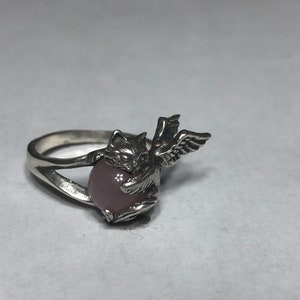May include: A silver ring with a pink stone and a cat with wings design.