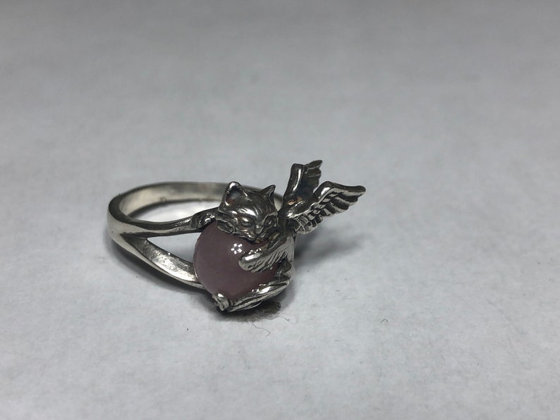 May include: A silver ring with a pink stone and a cat with wings design.