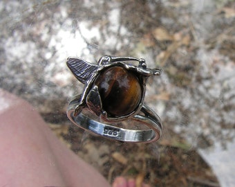 Könnte beinhalten: Ein silberner Ring mit einem Tigerauge-Stein, der in einem Kolibri-Design gefasst ist. Der Ring ist mit "925" auf der Band gekennzeichnet.