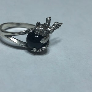 May include: A silver ring with a black gemstone and a winged cat design.