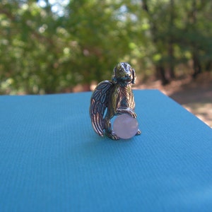 May include: A silver charm depicting a winged creature holding a pink gemstone. The charm is displayed on a blue surface.
