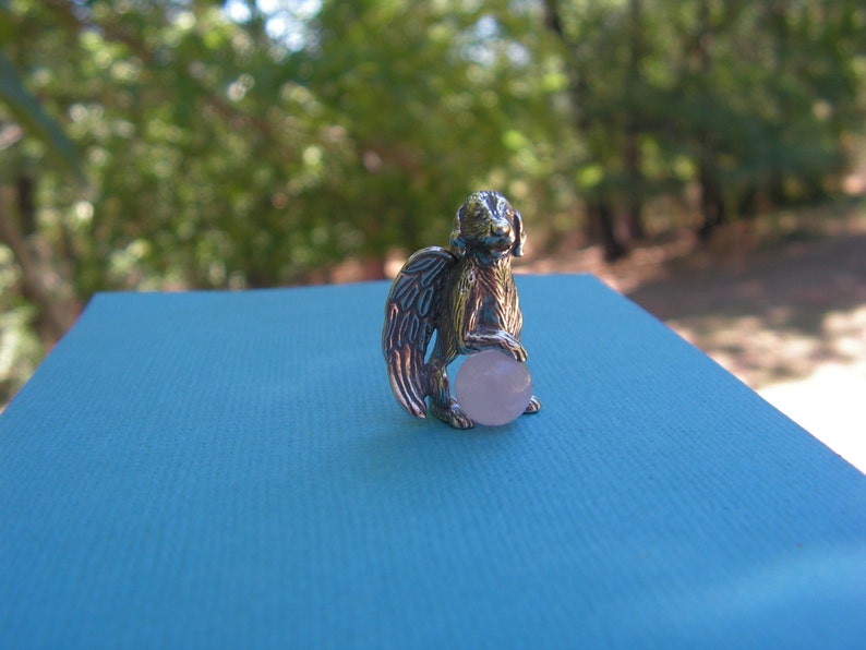May include: A silver charm depicting a winged creature holding a pink gemstone. The charm is displayed on a blue surface.