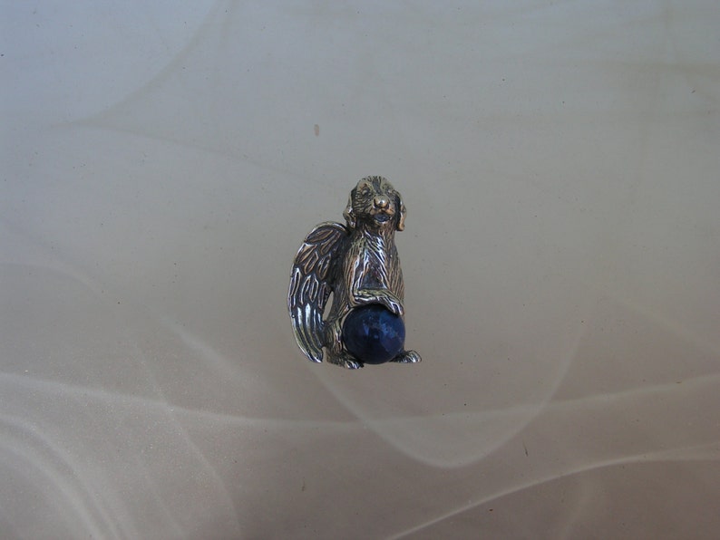 May include: A silver charm of a dog with wings holding a blue gemstone ball.