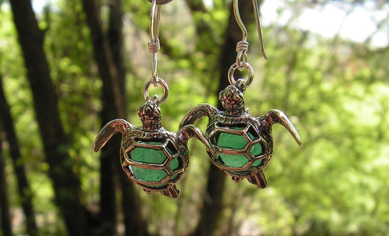 Pu&ograve; includere: Un paio di orecchini a forma di tartaruga in argento con accenti in vetro verde. Le tartarughe sono dettagliate con un design a guscio e hanno un gancio pendente.