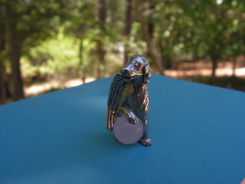 May include: A silver figurine of a dog holding a pink ball. The dog is standing on a blue surface.