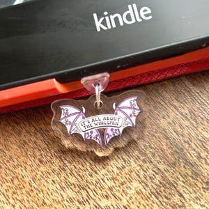 Puede incluir: Un lector de libros electrónicos Kindle negro con un borde rojo y la palabra "kindle" en blanco. Un dije acrílico transparente con un diseño de murciélago púrpura y el texto "IT'S ALL ABOUT THE WINGSPAN" está adjunto al dispositivo.