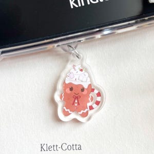 Könnte beinhalten: Ein klarer Acryl-Charm mit einem Lebkuchenmann-Design in einer Tasse. Der Charm hat rote, weiße und braune Farben. Das Wort "kindle" ist auf einem schwarzen Bildschirm zu sehen. Der Text "Klett-Cotta" ist ebenfalls sichtbar.