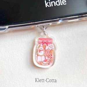 Puede incluir: Un charm de acrílico transparente con forma de tarro lleno de galletas navideñas, un muñeco de nieve y un bastón de caramelo. El charm cuelga de un lector Kindle. La palabra "kindle" es visible.