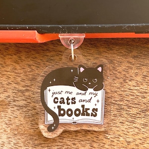 May include: A clear acrylic keyring with a black cat design. The cat is lying on a book, and the keyring has the text "just me and my cats and books". The cat has pink ears and is surrounded by stars.