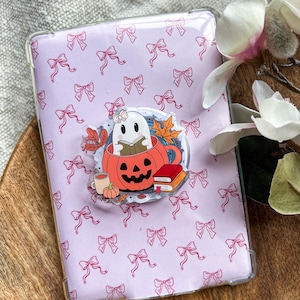 Op de afbeelding: Een rechthoekig transparant telefoonhoesje met een lichtroze achtergrond en een herhalend patroon van kleine roze strikjes. In het midden staat een ronde afbeelding van een wit spookje met een strikje in een pompoenmok.