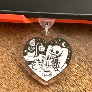 Puede incluir: Un llavero en forma de corazón con una ilustración en blanco y negro de un esqueleto leyendo en la cama con un gato, una lámpara y una taza de café. El fondo es una escena de noche estrellada. La palabra "kindle" es visible.