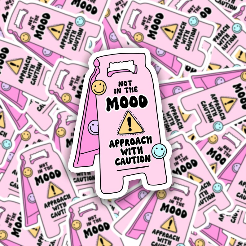 Op de afbeelding: Roze en witte waarschuwingsteken sticker met de tekst "Not in the Mood" en "Approach with Caution". Het bord is gevormd als een traditioneel waarschuwingsbord met een geel uitroepteken en een smiley.