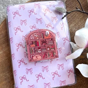 Può includere: Una custodia protettiva trasparente con un motivo a fiocco rosa, contenente un tablet con un design a fiocco abbinato. Una spilla decorativa a forma di libreria con libri, un gatto e un fantasma è attaccata al tablet.